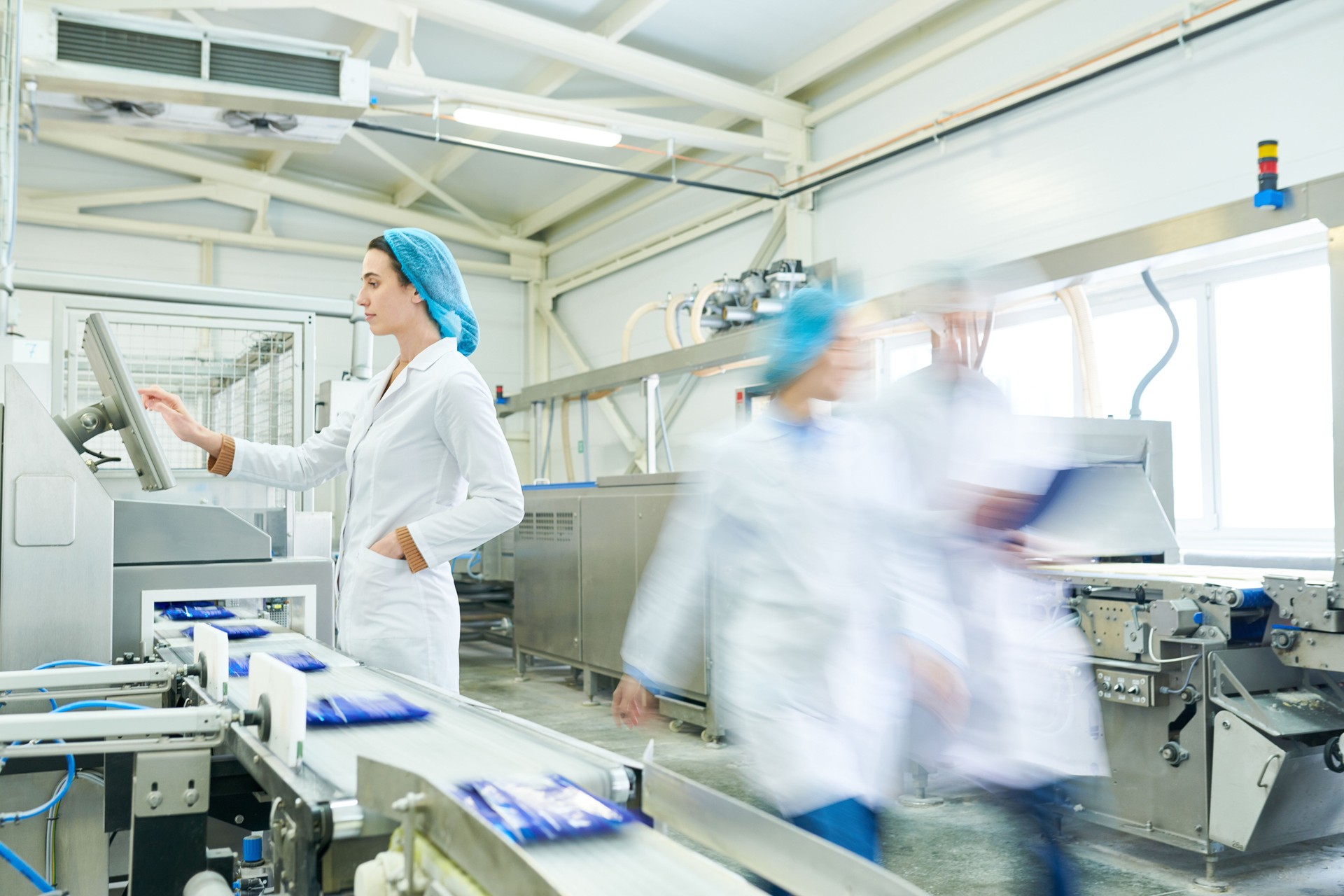 Busy female worker in sterile clothes choosing program on touch screen while operating manufacturing machine producing packaged food, blurred motion of technologists